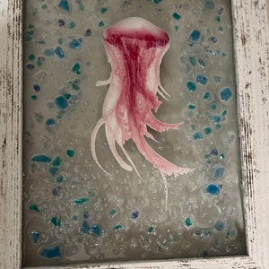 May include: A pink jellyfish encased in resin with blue and green glass pieces. The jellyfish is suspended in a clear resin with a white background.