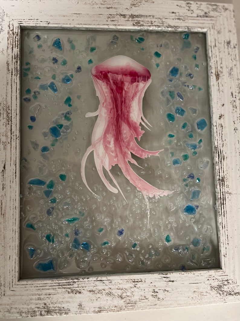 May include: A pink jellyfish encased in resin with blue and green glass pieces. The jellyfish is suspended in a clear resin with a white background.