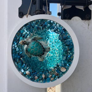 May include: A white circular frame with a resin-filled center featuring a sea turtle swimming in a turquoise blue ocean with white seashells. The resin is filled with blue and white glass pieces.