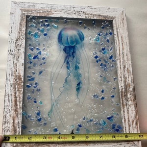 May include: A framed artwork featuring a blue and white jellyfish with a translucent body and long tentacles. The jellyfish is surrounded by small blue glass pieces, creating a textured and dimensional effect. The frame is white with a distressed finish.