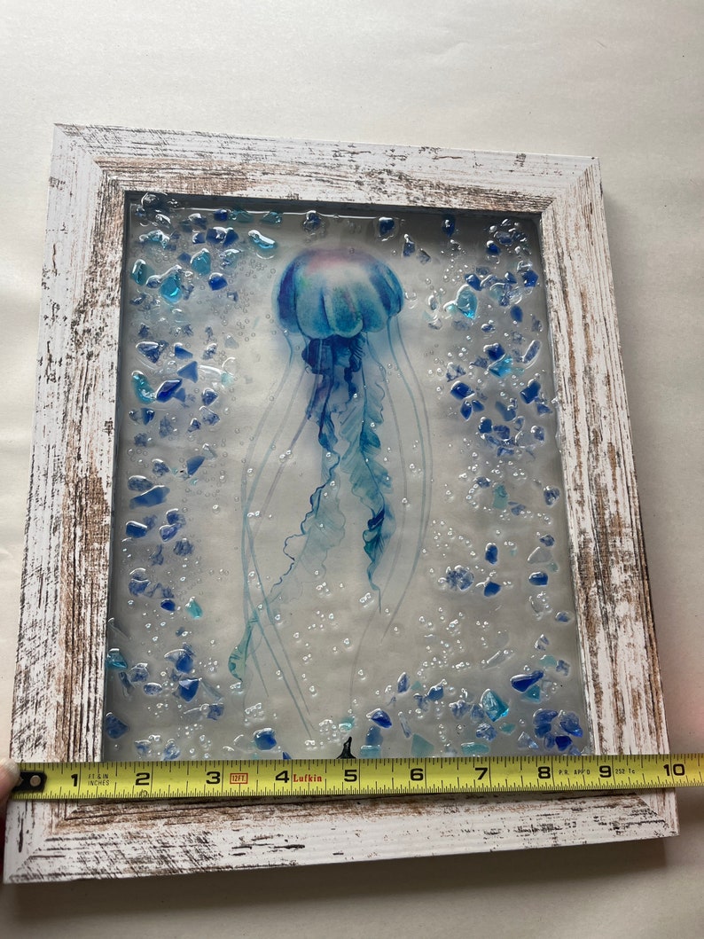 May include: A framed artwork featuring a blue and white jellyfish with a translucent body and long tentacles. The jellyfish is surrounded by small blue glass pieces, creating a textured and dimensional effect. The frame is white with a distressed finish.