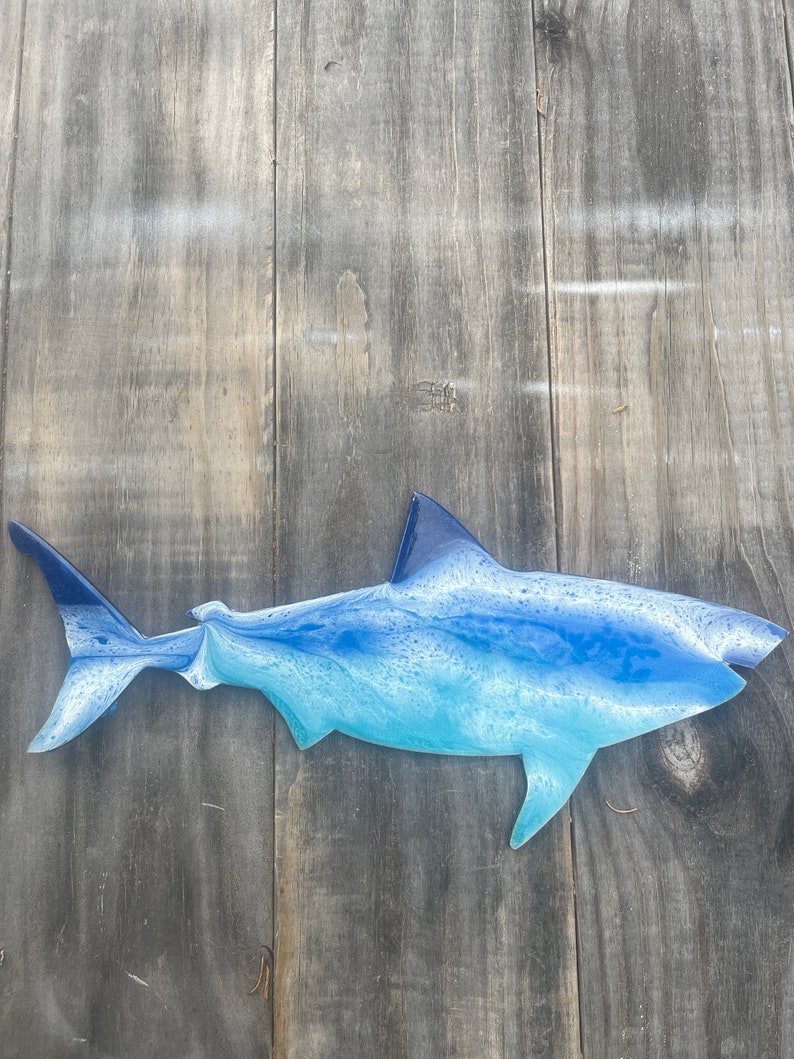 May include: A blue and white resin shark wall sculpture. The shark is in a side profile with its mouth closed. The sculpture is mounted on a wooden background.