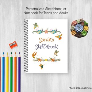 May include: A white spiral-bound sketchbook with the text "Sarah's Sketchbook" and hummingbird illustrations. The cover is decorated with floral and hummingbird designs. A set of colored pencils and a pencil sharpener are on the left.