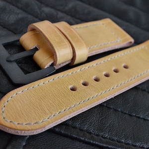 Puede incluir: Una correa de reloj de cuero marrón claro con hebilla negra y costuras blancas. La correa tiene varios agujeros para ajustar el tamaño.