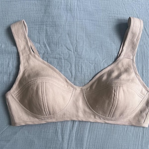 May include: A beige bralette featuring wide shoulder straps and a deep scoop neckline. The cups are constructed with visible vertical seam detailing, providing a structured shape. The garment is crafted from a solid, neutral-toned fabric with a matte finish.