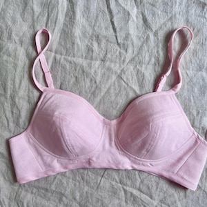 May include: A light pink brassiere featuring moulded cups with visible stitched paneling, adjustable shoulder straps, and a wide underband. The garment is constructed from a solid-coloured fabric with a subtle texture, showcasing a classic design.