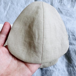 May include: A beige, unstructured hat made of a textured fabric. The hat is held in a hand, showcasing its shape and material. The hat appears to be a casual accessory.