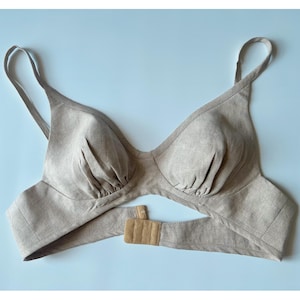 Könnte beinhalten: Ein beigefarbenes Bralette mit gerafften Stoffkörbchen, dünnen Schulterträgern und einem breiten Unterbrustband, das am Rücken mit einem Hakenverschluss geschlossen wird. Das Kleidungsstück besteht aus strukturiertem, neutralfarbenem Stoff mit sichtbaren Nähten.