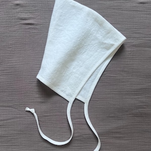 Puede incluir: Un gorro blanco hecho de una tela texturizada con una corona puntiaguda y un borde frontal redondeado. La prenda cuenta con dos cintas de tela largas y delgadas que se extienden desde la base, cada una con un pequeño nudo en el extremo.
