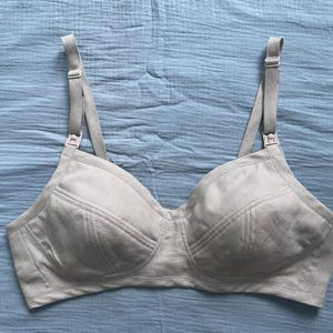 May include: A beige nursing bra featuring moulded cups with visible seam detailing, adjustable shoulder straps, and plastic nursing clips at the top of each cup. The garment is constructed from a solid-coloured fabric with a textured appearance.