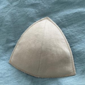May include: A beige, triangular-shaped fabric item with a stitched border, resting on a light blue textile surface. The item has a seam running down the center, creating two symmetrical halves. The fabric appears to be linen or a similar material.