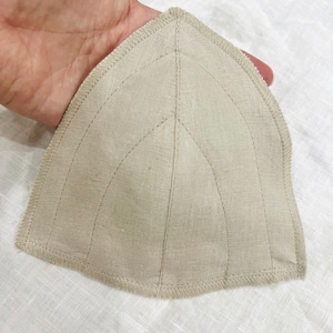 以下が含まれることがあります： ベージュ色の三角形の布製アイテムで、ステッチのディテールが施されています。アイテムは丸みを帯びたエッジと、ステッチによって作られた葉のようなデザインが特徴です。生地はリネンまたは同様の天然素材のようです。アイテムは手で持たれています。