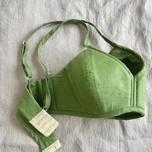 May include: A grass green bralette featuring structured cups with vertical seams, adjustable shoulder straps, and a beige hook-and-eye closure at the back. The garment is crafted from a textured fabric and displays a solid, vibrant green colour throughout.