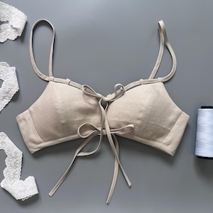 Op de afbeelding: Een beige bralette met dunne schouderbandjes, een striksluiting aan de voorzijde met lange stoffen linten en een gestructureerd lijfje. Een strook witte kanten band met bloemmotief en een klosje wit naaigaren liggen naast het kledingstuk.