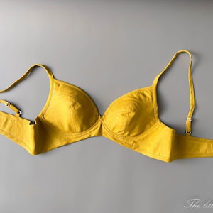 May include: A mustard yellow bra with adjustable straps and a hook-and-eye closure. The bra is made of a textured fabric and is set against a grey background. The Little Heidi is written in cursive at the bottom.