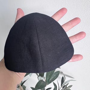 May include: A black, triangular-shaped head covering, possibly a kippah or yarmulke, made of a textured fabric. The item is held in a hand against a white background with green foliage.
