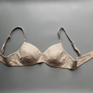 May include: A beige bra with adjustable straps and a hook-and-eye fastening. The bra features a classic design with moulded cups and underwire support. The fabric appears to be a soft, natural material.