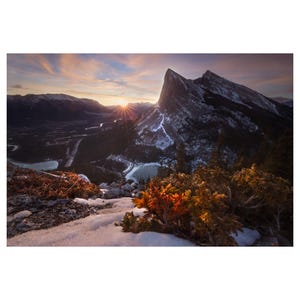 May include: A panoramic view of a snow-capped mountain range at sunrise. The sun is shining brightly behind the mountain peaks, casting a golden glow over the landscape. The foreground features a small patch of snow-covered ground with a few evergreen bushes.