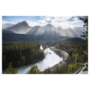 May include: Scenic landscape photograph featuring a train winding through a lush green forest alongside a river. Mountains rise in the background, with sunlight streaming through the clouds, creating a dramatic and picturesque scene.