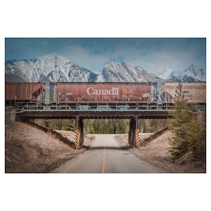 May include: A rusty freight train with a boxcar labeled "Canada" travels over a bridge with a paved road underneath. The train is traveling through a mountain pass with snow-capped peaks in the background.