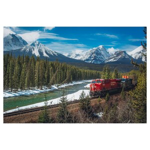 May include: A red train travels through a snowy mountain valley. The train is traveling on a track that runs along the edge of a river. The mountains are covered in snow and the sky is blue.