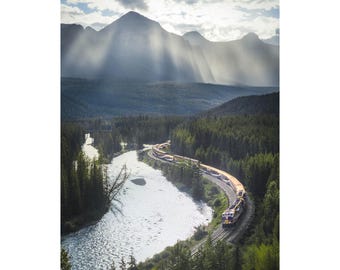 May include: Scenic landscape photograph featuring a train winding along a river through a lush forest. Mountains rise in the background, with sunlight streaming through the clouds. The train is yellow and blue.