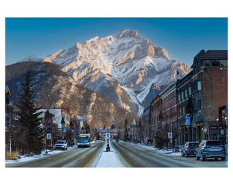 May include: A blue bus drives down a snow-covered street in a town with a mountain in the background. The buildings are brick and have snow on their roofs. There are trees on either side of the street.