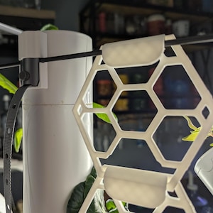 May include: White plastic plant support with a hexagonal grid design. The support is attached to a white pipe with a black strap.