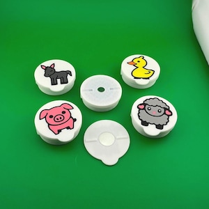 May include: A set of six circular white plastic covers, four featuring colorful cartoon animal illustrations of a donkey, a yellow duck, a pink pig, and a grey sheep. Two covers are plain white with a central circular magnetic component.