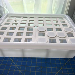May include: A rectangular white plastic storage bin featuring a removable insert tray. The tray has a grid pattern with square openings and several circular solid white discs integrated into the top surface, providing a structured organization system.