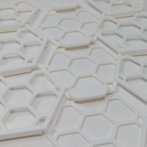 May include: A close-up of white plastic honeycomb-shaped tiles stacked on top of each other. The tiles have a raised edge around the perimeter.