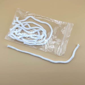May include: A clear plastic bag containing several white, braided cords. One cord is separated from the others. The cords are white and appear to be made of a soft, flexible material. The background is a solid, light tan color.