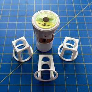 May include: A white AeroGarden seed pod kit for mint, featuring a central cylindrical pod with a green label reading Mint (Sprouts in 6-10 days) Medium, surrounded by three white plastic support baskets with arched openings and circular bases.