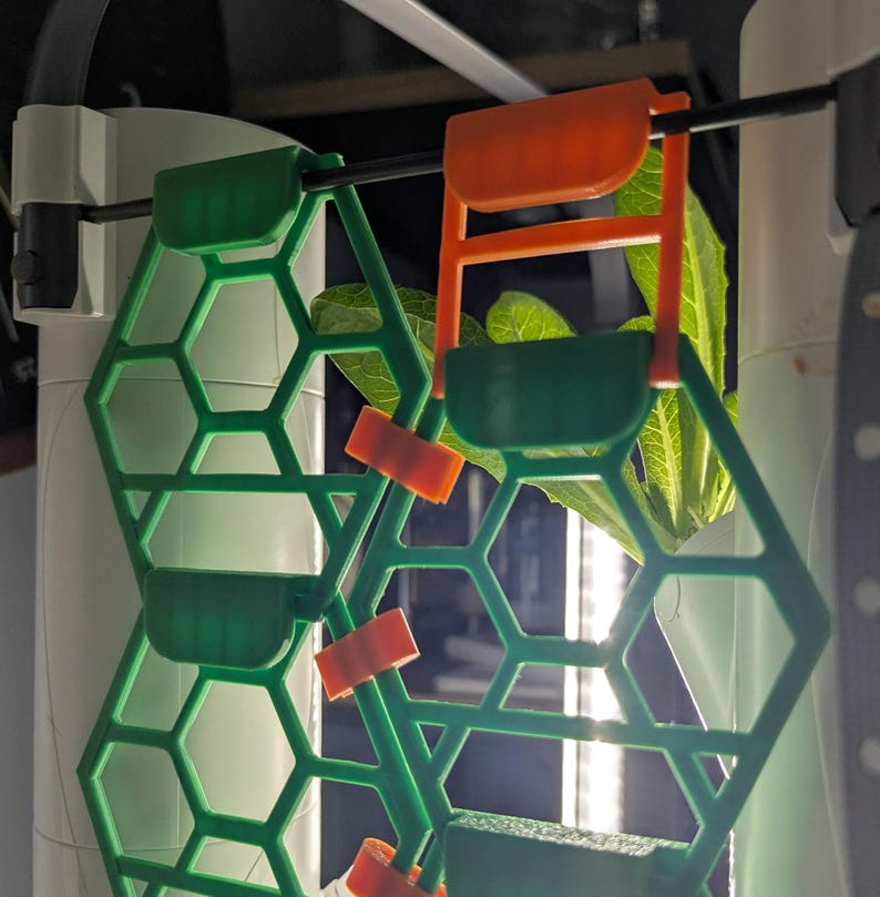 May include: Green and orange plastic plant supports with a honeycomb design. The supports are attached to a white pole and are holding up a green plant.