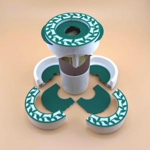 May include: A white and green hydroponic system with a circular top and base, featuring a green leaf pattern. The system includes a central white cylinder and several green and white curved pieces. The components are arranged on a light orange surface.