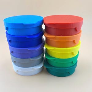 May include: Stacks of colorful, round plastic containers in shades of blue, red, orange, yellow, and green. Each container has a lid with a small tab for easy opening. The containers are stacked neatly, showcasing a variety of vibrant colors.