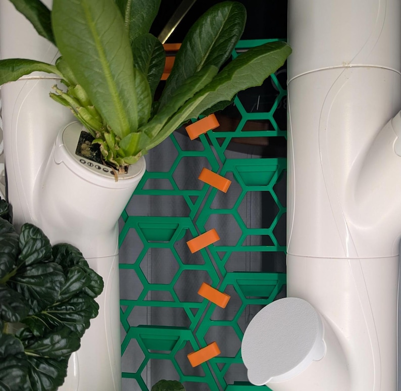 May include: A white vertical hydroponic gardening system with green plants growing in white pots. The system has a green honeycomb-shaped panel with orange clips.