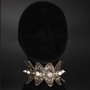 May include: A wide, ornate gold-tone metal choker necklace featuring intricate filigree patterns and small, round white pearls set at intervals along the band. The necklace has a structured, scalloped edge design that encircles the neck.