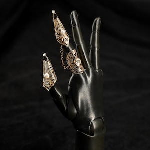May include: Ornate gold-colored finger rings with intricate filigree designs and sparkling clear gemstones are displayed on a black hand. The rings include a chain connecting two of them, adding a unique touch to the jewelry.