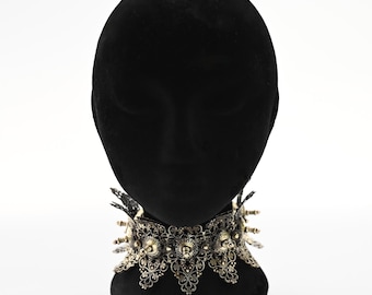 May include: A wide, ornate choker necklace featuring intricate, dark metallic filigree patterns. The design includes small, protruding spikes and decorative metal elements arranged in a repeating, scalloped edge formation around the neck.