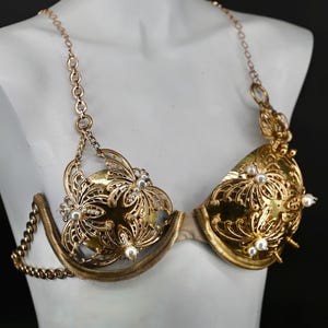 May include: A gold-colored, ornate bra with intricate floral and filigree designs. The bra features pearl accents and chain straps. The garment is displayed on a white mannequin against a black background.
