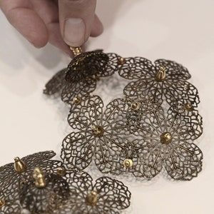 May include: A delicate gold-toned metal jewelry piece featuring a cluster of intricate, openwork flower designs. The flowers are connected to form a larger, geometric shape.