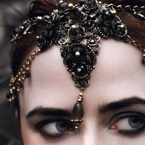 May include: A gold and black jeweled headpiece with a dangling teardrop-shaped pendant. The headpiece is adorned with intricate floral designs and black beads.