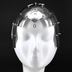 May include: A silver headpiece with a geometric design and gold accents. The headpiece has a small loop hanging from the centre.