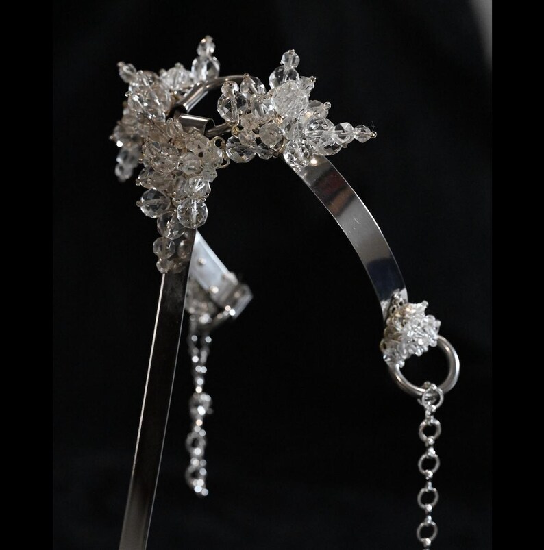 May include: A silver-toned headpiece with clear, faceted crystal accents. The design features a curved band and vertical supports, with dangling chain and ring details. The crystals are clustered at key points, creating a sparkling effect against a dark background.