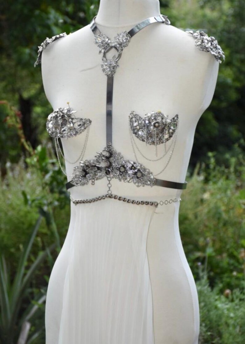 May include: A silver-toned body harness with ornate crystal embellishments. The harness features shoulder accents, a chest piece, and a waist chain. The design includes dangling chains and is displayed on a white mannequin against a blurred green background.