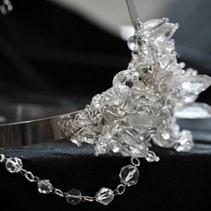 May include: A silver tiara with a cluster of clear, faceted crystals. A delicate chain of clear beads hangs from the side. The tiara is set against a dark background, highlighting the sparkle of the crystals.