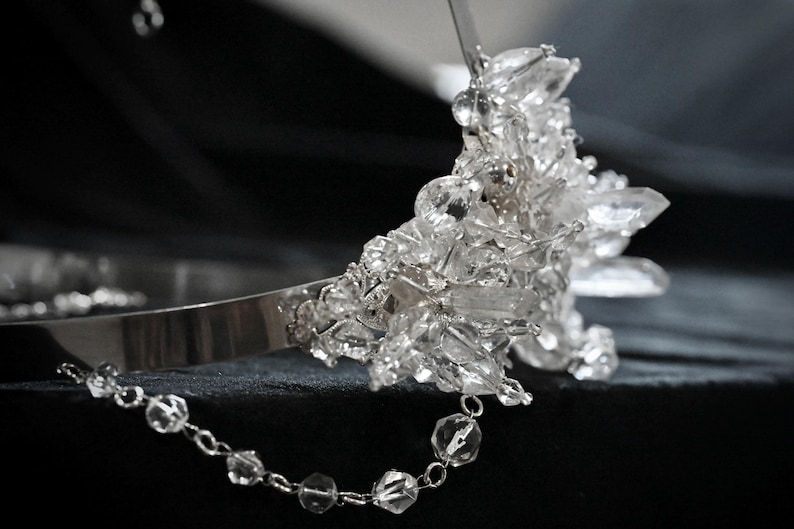May include: A silver tiara with a cluster of clear, faceted crystals. A delicate chain of clear beads hangs from the side. The tiara is set against a dark background, highlighting the sparkle of the crystals.