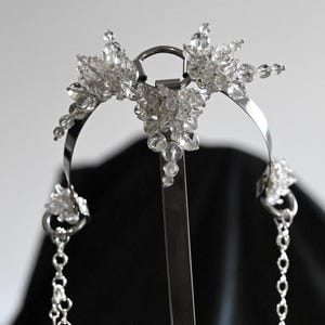 May include: A silver-toned headpiece with clear crystal accents. The headpiece features a central floral design and dangling chains. The crystals are clustered in floral shapes, adding sparkle. The design is symmetrical and elegant.