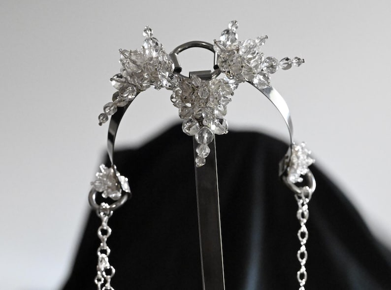 May include: A silver-toned headpiece with clear crystal accents. The headpiece features a central floral design and dangling chains. The crystals are clustered in floral shapes, adding sparkle. The design is symmetrical and elegant.